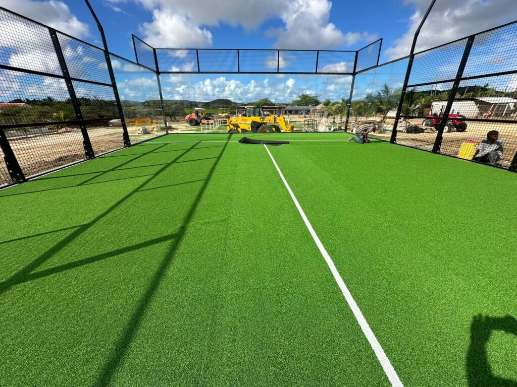 Installation of an LDK classic panoramic glass padel court in Bonaire, showing the bright green artificial turf and construction equipment in the background.