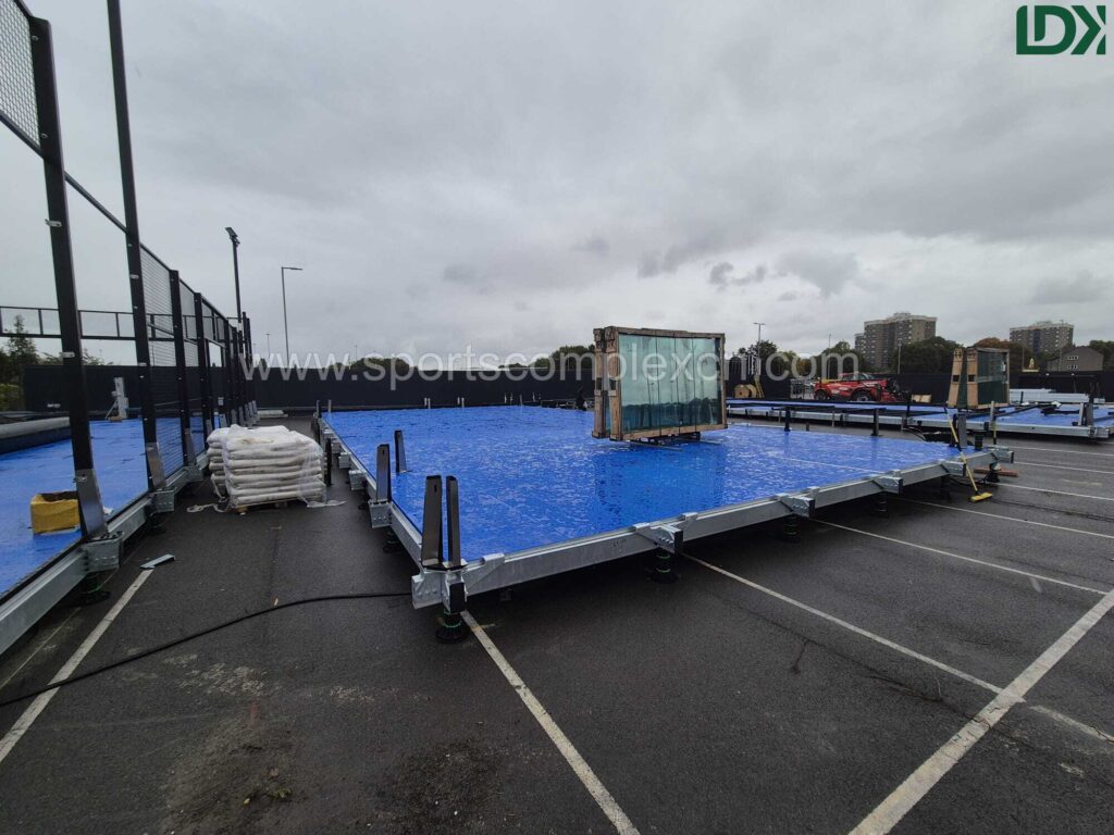Partially installed mobile padel court with blue artificial turf and glass panels being set up on an asphalt surface.