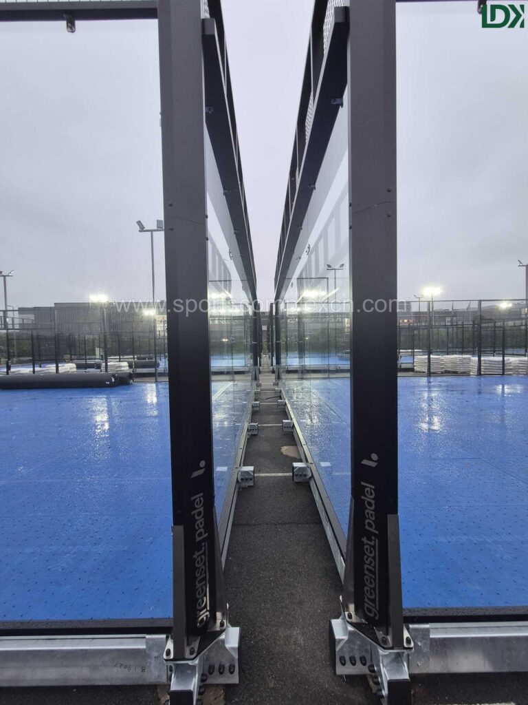 Ground-level view looking down the narrow gap between two parallel mobile padel courts, showing their exterior structural supports.