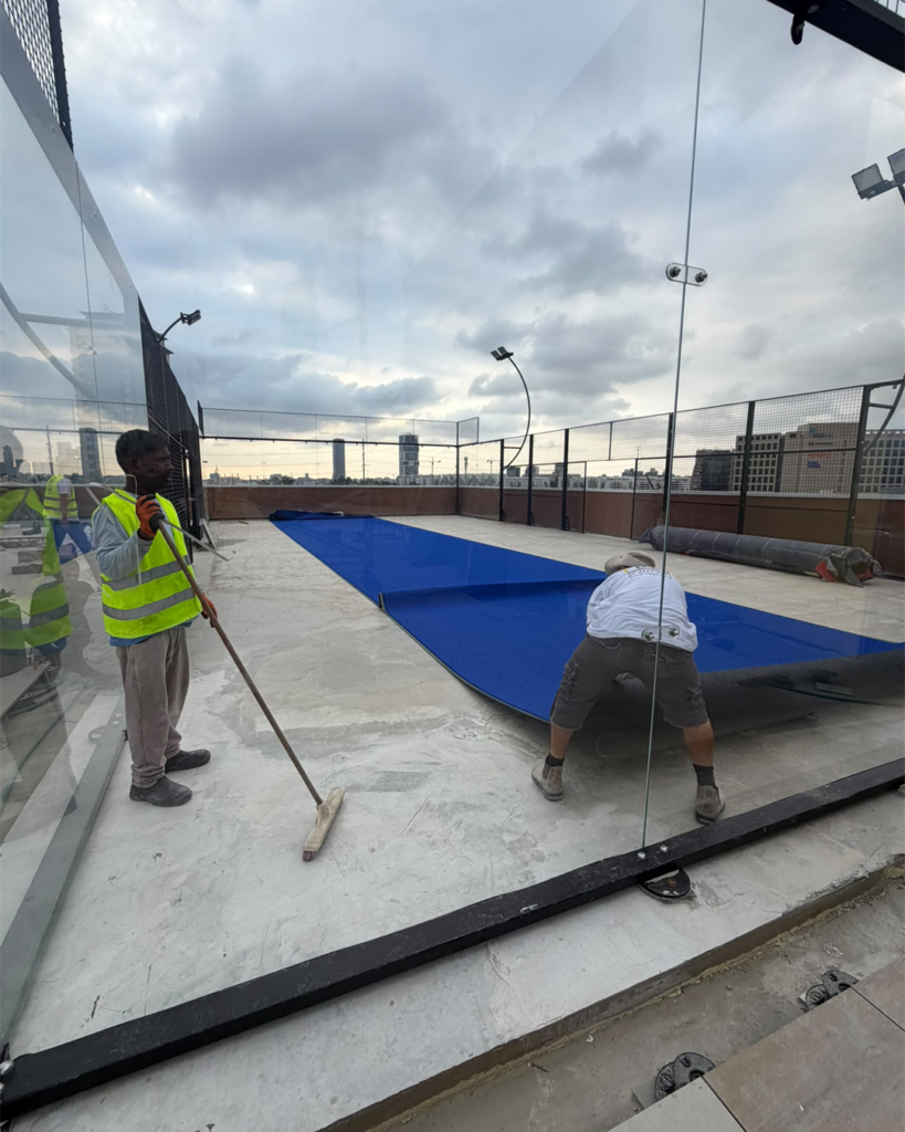 Installers align blue artificial turf across the padel court floor.