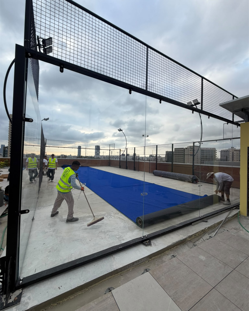 Installers fitting blue artificial turf inside a glass-enclosed panoramic padel court.